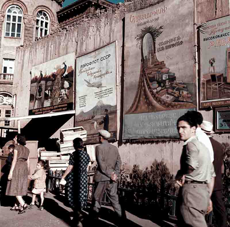 Historical photograph of people walking by a  a wall with multiple posters and advertisements in Armenia during the Soviet era.