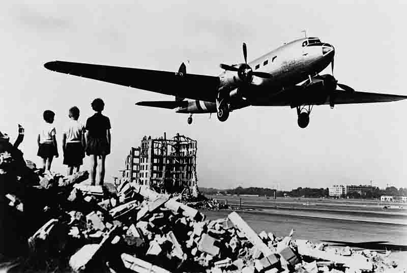 A historic black-and-white photo of a Douglas C-47 'Raisin Bomber' transport plane landing at Berlin's Tempelhof Airport during the Berlin Airlift (1948–1949), while three children watch from atop a massive pile of war rubble.