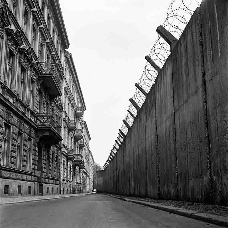 A black-and-white, low-angle photograph capturing a stark perspective of the Berlin Wall as it runs parallel to a row of residential buildings.