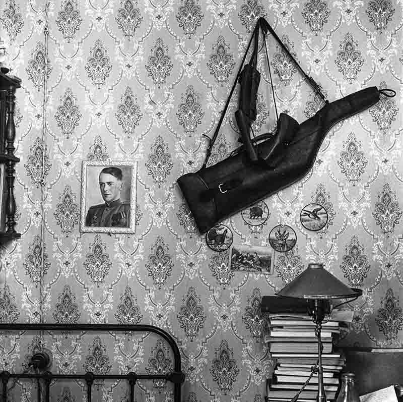 A black and white image of a bedroom featuring a bed, scattered books, a portrait photo and a gun on the wall.