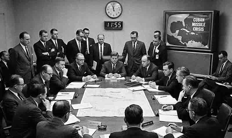 A black and white photograph depicting a tense meeting in a conference room during the Cuban Missile Crisis.