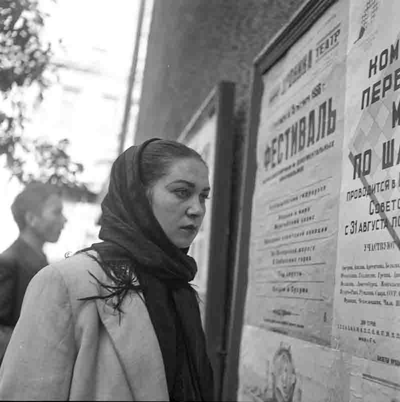 A Soviet woman with a headscarf stands in front of a poster with a message looking thoughtfully at the depicted object.