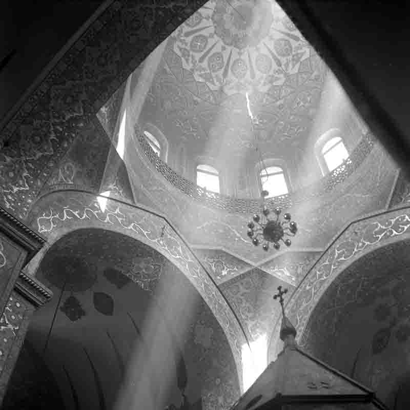 Etchmiadzin Cathedral Dome A high-angle black and white shot of a Etchmiadzin Cathedral Dome, looking up from below. Ornate, decorated arches lead the eye up to the beautifully patterned dome, which has a small chandelier hanging from its center.