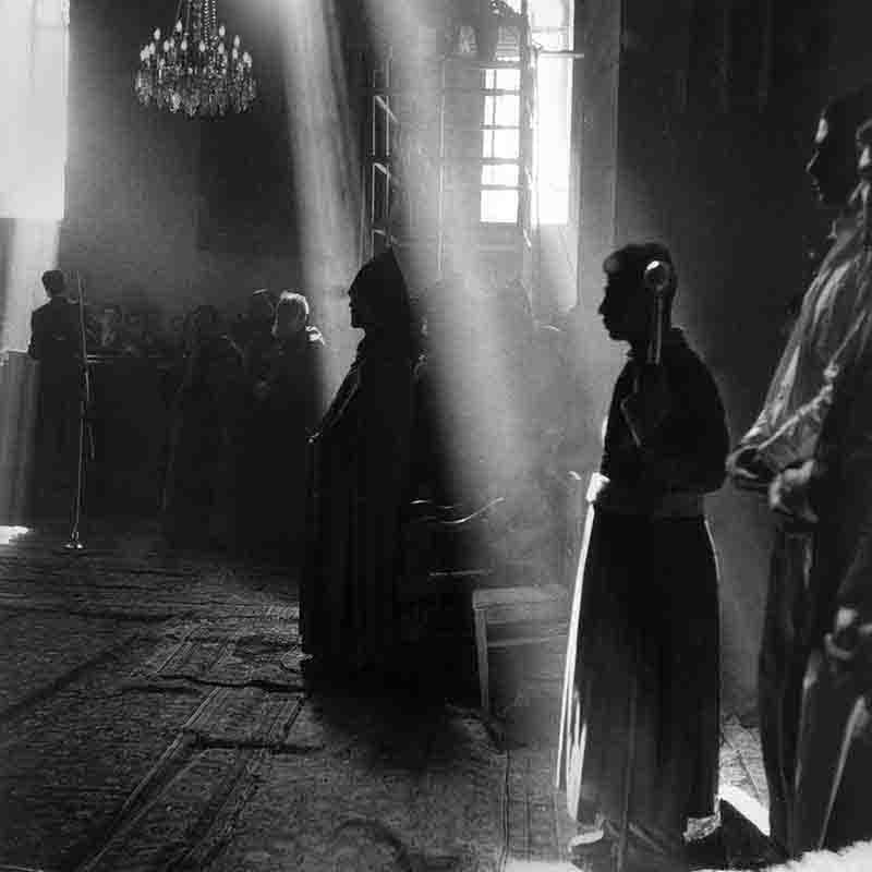Etchmiadzin Cathedral: Seat of the Armenian Apostolic Church Sunlight streams through the windows of a dimly lit Etchmiadzin cathedral, illuminating dust motes in the air and casting dramatic rays onto the carpeted floor. Silhouetted figures, some in religious attire, stand within the space, their forms partially obscured by the light and shadow. The overall atmosphere is solemn and spiritual.