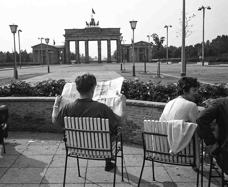 A poignant black-and-white study of daily life in divided Berlin. In the foreground, a man sits in a slatted cafe chair, his attention fully absorbed by a newspaper, while the Brandenburg Gate looms in the background.