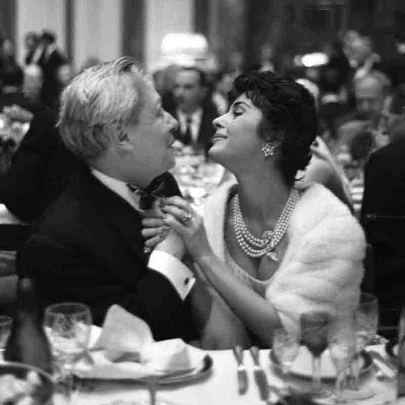 A monochrome photograph showcasing a man in a tuxedo sits at a table with a woman, both engaged in conversation, creating an elegant atmosphere.