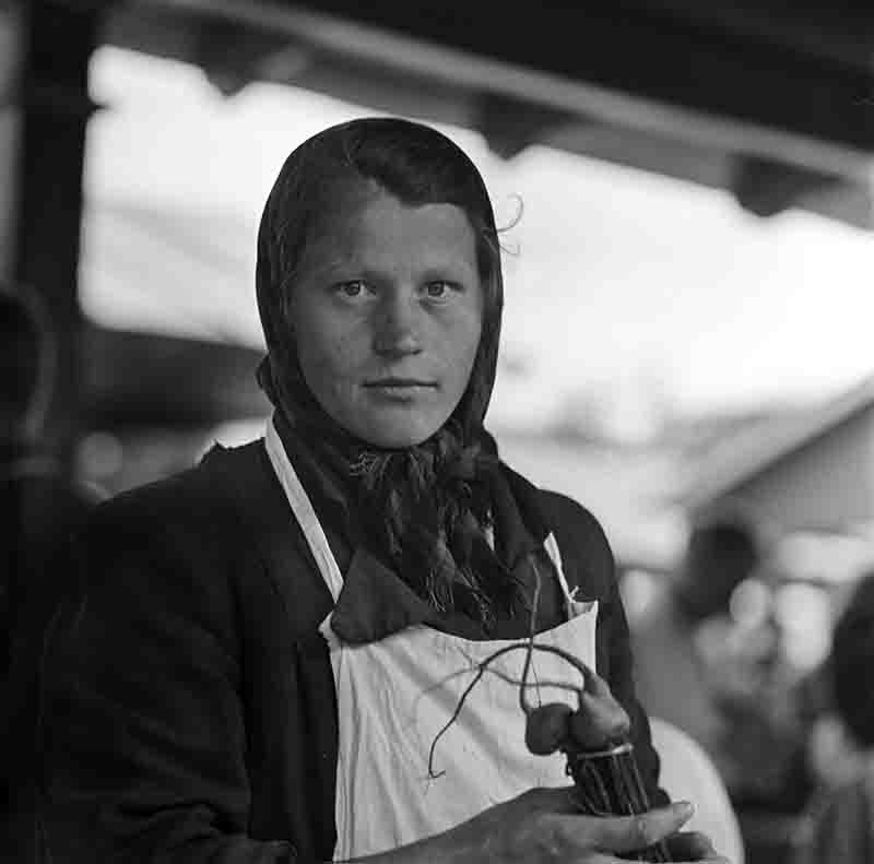 A monochrome photograph showcasing a woman in a headscarf stands at a market displaying vegetables