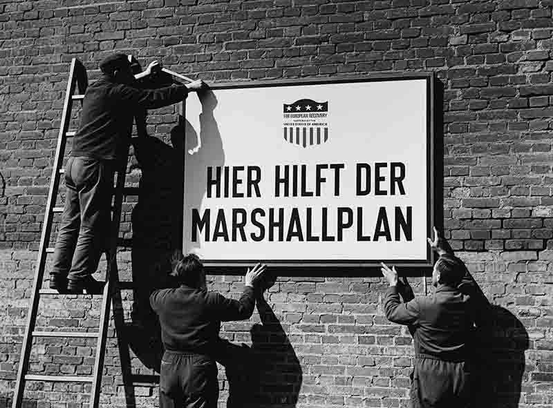 A black and white photograph showing four men mounting a large rectangular sign onto a dark brick wall. One man stands on a wooden ladder on the left, reaching toward the top of the sign, while three others support it from the bottom and sides.