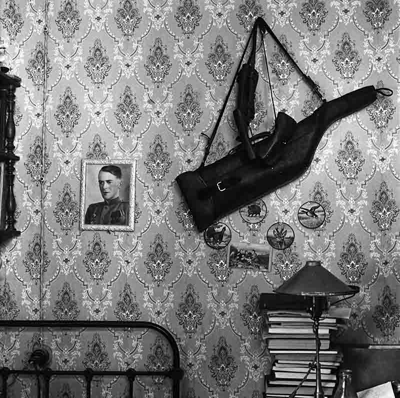 May 9 Victory Day Black and white photo of a fallen Soviet soldier's bedroom with a bed, books on a shelf and his portrait and rifle in honorific display on the wall