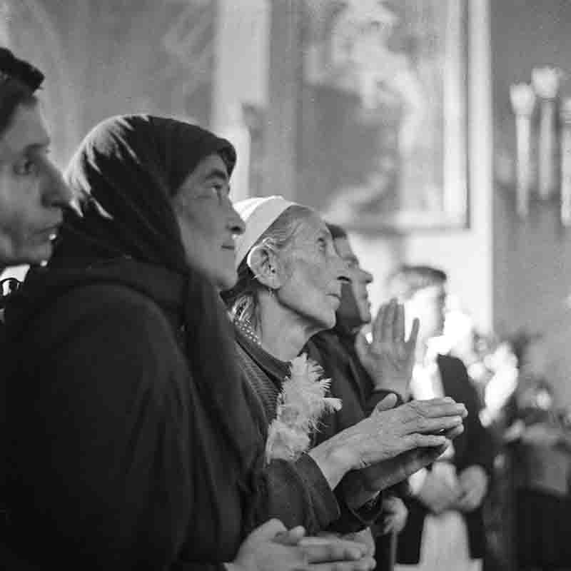 Black and white photograph captures a row of worshipers standing and praying inside a Russian Orthodox church.