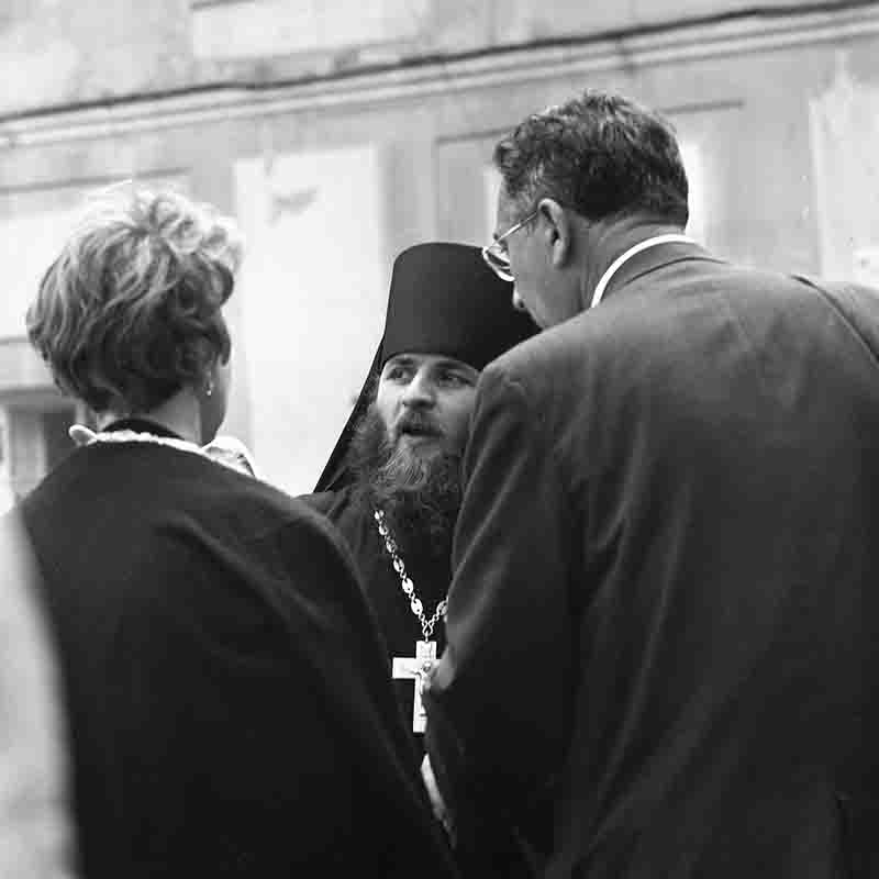 A monochrome photograph showing a Russian Orthodox Church Priest conversing with a couple, highlighting their interaction in a candid moment.