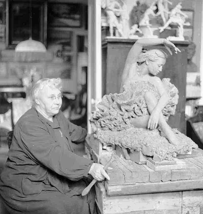 A historical black and white photograph showing Alexandrovna Yanson-Manizer engaged in sculpting, surrounded by her artistic tools.