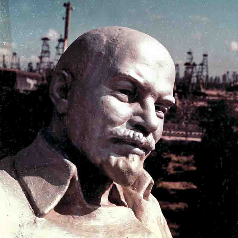 Soviet Azerbaijan Lenin Bust A close-up, color-tinted photograph of a metallic or stone bust of Vladimir Lenin. The statue shows him with a bald head, a goatee, and a serious, downward gaze, typical of his depictions. In the background, out of focus, a field of Soviet-era Azerbaijan oil derricks or drilling rigs extends toward the horizon under a pale blue sky.