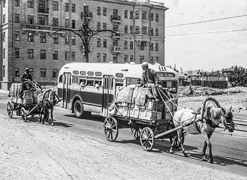 Street life in Moscow, 1956 A black and white photograph from 1956 Moscow. The image shows a street scene with two horse-drawn carts and a bus. The bus is in the center of the frame, with the horse-drawn carts on either side. A man is riding on each cart. The background shows a large building and some trees.