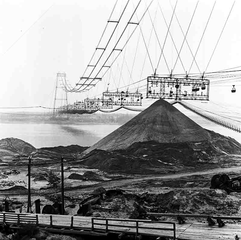 A black and white photo shows a large construction site. In the foreground, a wooden walkway leads towards a massive, conical pile of earth or sand. Above the pile and extending into the distance are complex structures of suspended cables and platforms. These cables stretch across a wide body of water, and on the far side, a section of a suspension bridge is visible.