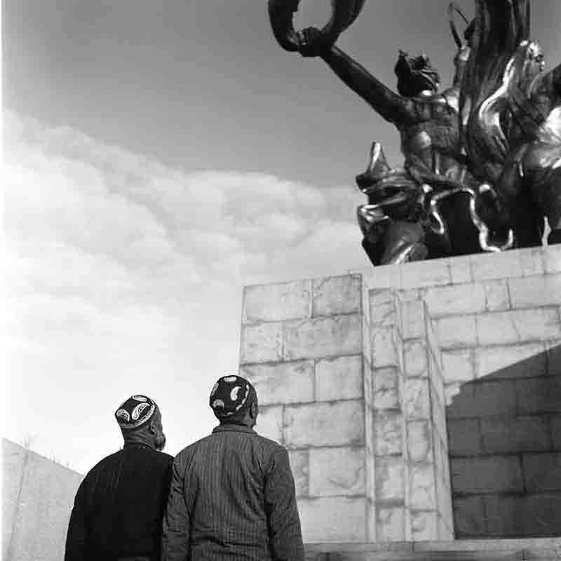 A monochrome photograph showcasing visitors from Uzbekistan stand before the Worker and Kolkhoz Woman statue, capturing a moment of respect and historical significance.