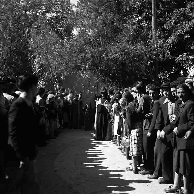 Vazgen I wearing a dark, hooded robe and a pointed hood . Behind him are other figures, some holding ceremonial banners or religious items, moving along the path. A crowd of people lines a curved path, looking toward the center of the image.