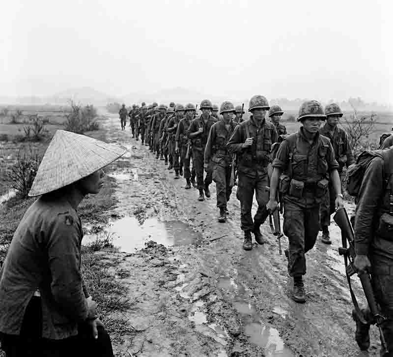 A black and white historical Vietnam war photograph shows a long column of soldiers marching down a muddy, rutted dirt road through a rural landscape. In the left foreground, a person wearing a traditional conical hat sits and watches the soldiers pass.