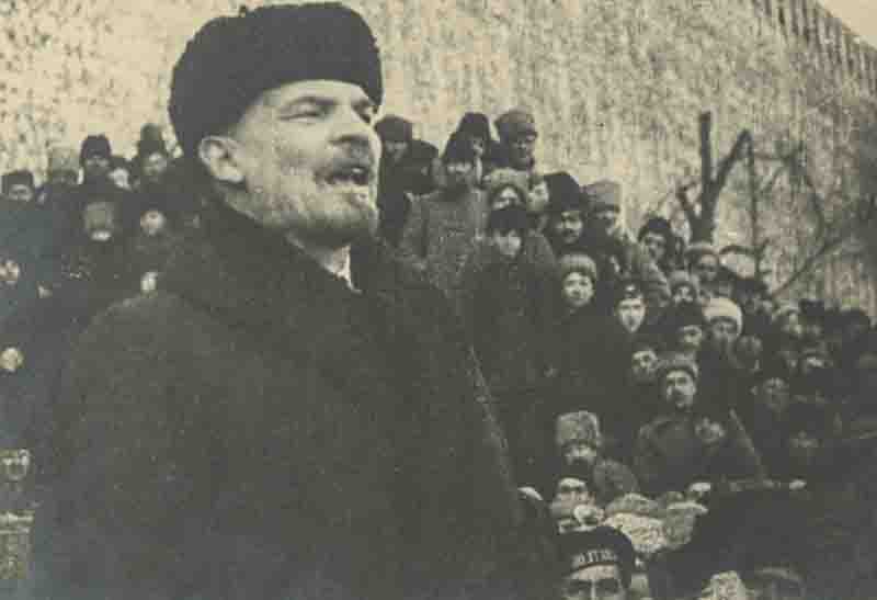 Black and white historical photograph of Vladimir Lenin wearing a dark winter coat and fur hat, speaking passionately to a gathered crowd outdoor near the Kremlin wall in Moscow.