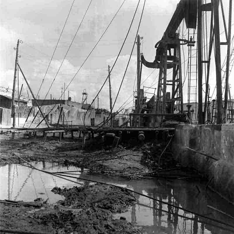 Black and white photo showing an old water pump in a backyard on an oil field in Baku, in the background a washing line with suspended laundry.