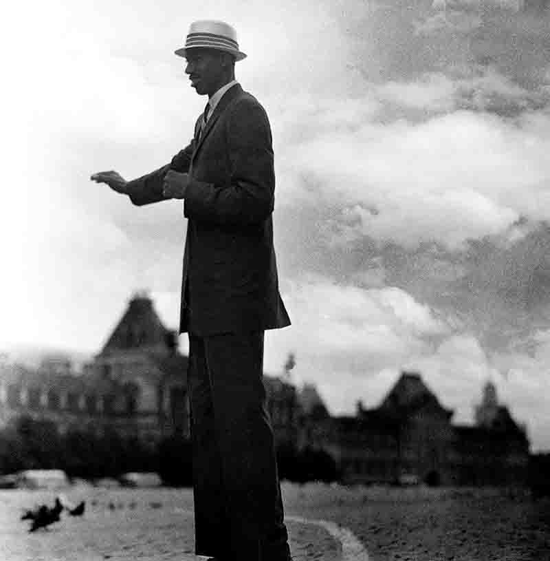 Wilt Chamberlain in Moscow A black-and-white, low-angle, full-body shot of Wilt Chamberlain standing on Moscow's Red Square. He's wearing a dark suit, tie, and a light-colored fedora hat. His right arm is outstretched, and his left hand is at his side. In the background, there's a cloudy sky and blurred buildings.
