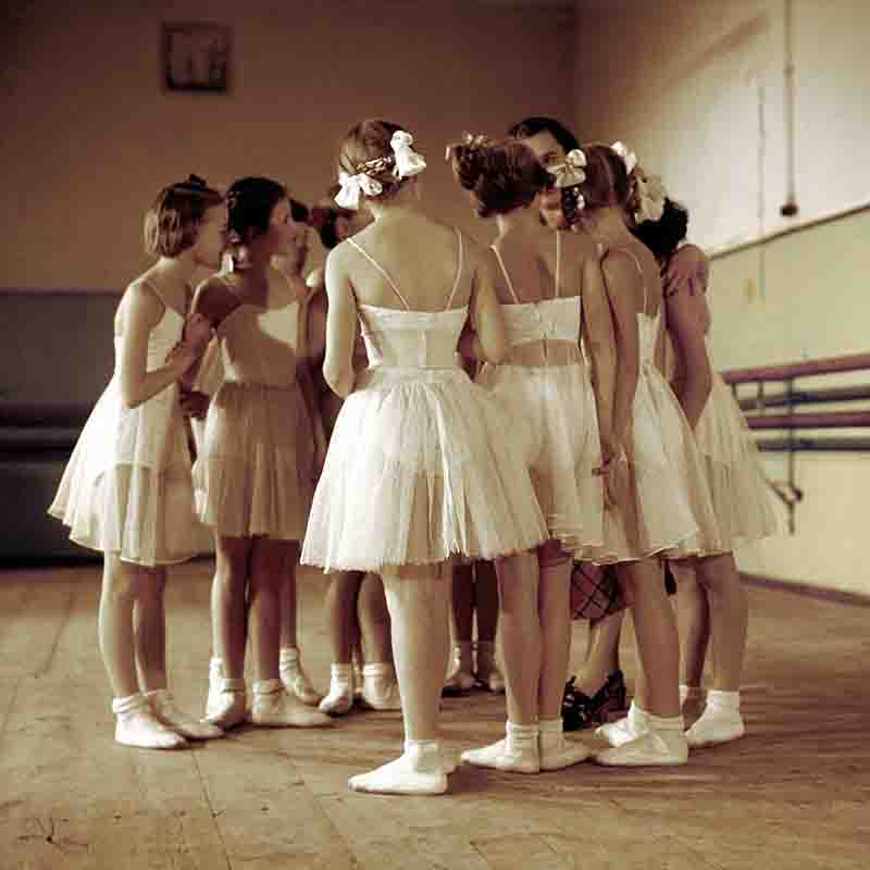 A group of young ballet dancers in the Bolshoi Theatre standing in a tight circle on a wooden dance floor, listening to an instructor, who is partially visible in the center.